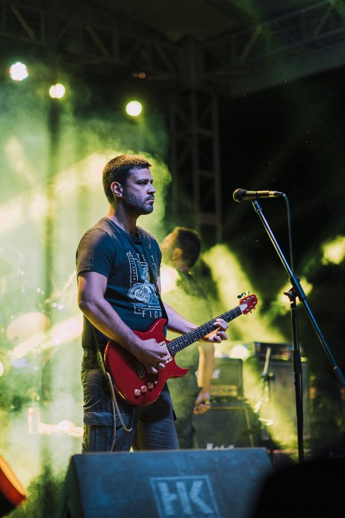Gitarzysta grający na czerwonej gitarze przy mikrofonie, w tle zielone światło i dym na scenie